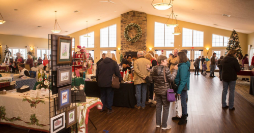 A festive indoor market with people browsing tables of holiday crafts and decorations, surrounded by seasonal decor.
