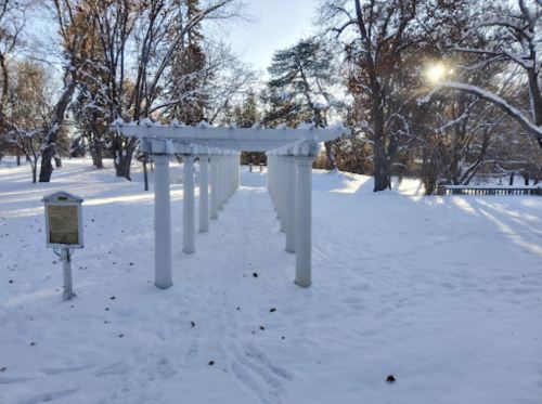 A snowy park scene featuring a white pergola surrounded by trees and sunlight peeking through the branches.
