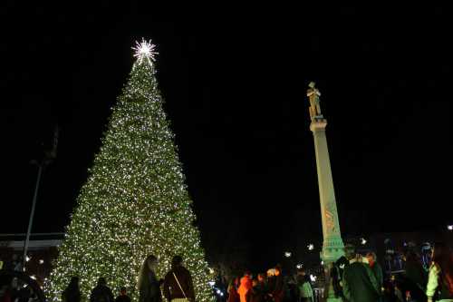 A large, illuminated Christmas tree stands tall at night, with people gathered around a nearby monument.