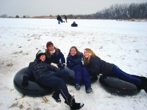 Four friends relax on snow tubes in a snowy landscape, with others sledding in the background.