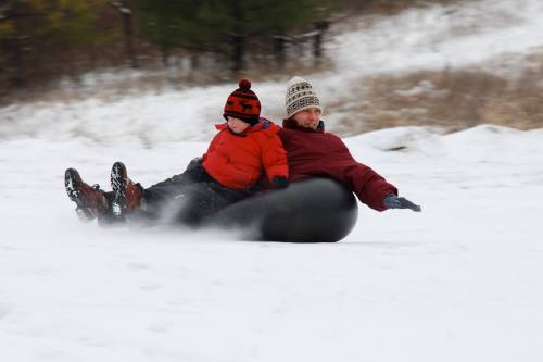 A child and an adult slide down a snowy hill on a black inner tube, both smiling and bundled up in winter clothing.