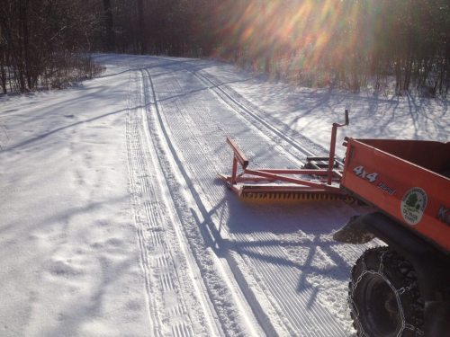 A snow-covered path being groomed by a snowcat, with sunlight shining through trees in the background.