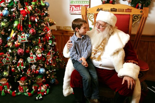 A young boy smiles at Santa Claus, sitting together by a decorated Christmas tree.