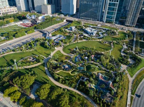 Aerial view of a lush urban park with pathways, greenery, and wind turbines, surrounded by city skyscrapers.