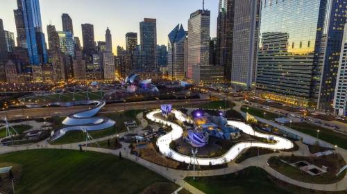 Aerial view of a city park with modern sculptures, pathways, and skyscrapers in the background during sunset.