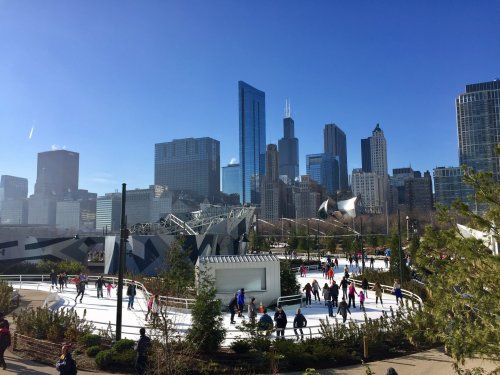 A bustling ice skating rink in a city park, surrounded by skyscrapers under a clear blue sky.