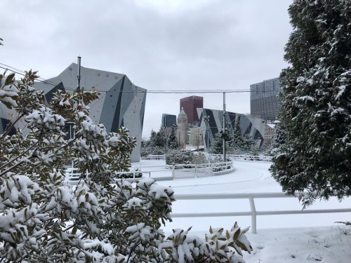 Snow-covered landscape featuring modern buildings and a cloudy sky, with greenery in the foreground.