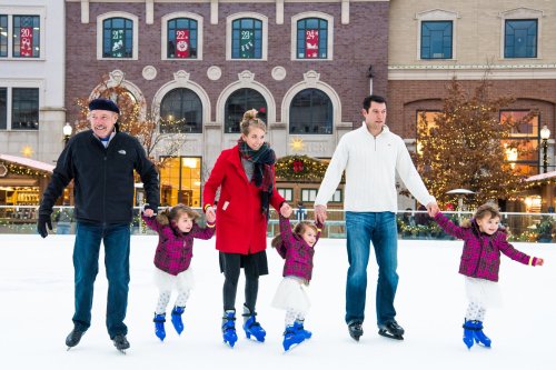 A family ice skating together, holding hands, with three young girls in matching outfits and festive decorations in the background.