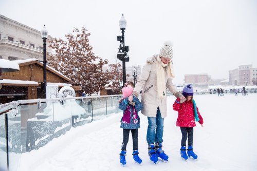 A woman ice skating with two children in a snowy outdoor rink, surrounded by winter scenery and buildings.