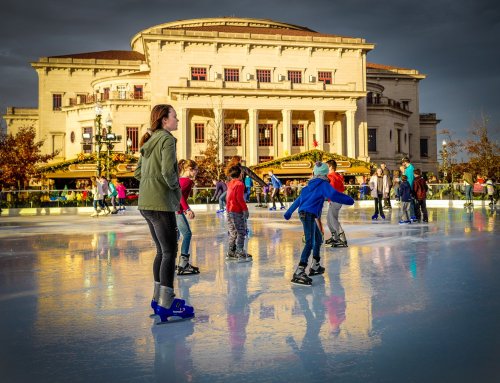 A group of children and adults ice skating on a rink in front of a grand building, with cloudy skies above.