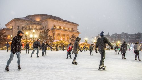 People ice skating in a snowy outdoor rink, surrounded by festive lights and a historic building in the background.