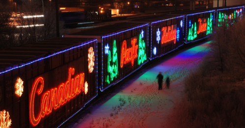 Colorful holiday lights illuminate train cars, spelling "Canadian" and featuring festive snowflakes and trees in a snowy landscape.