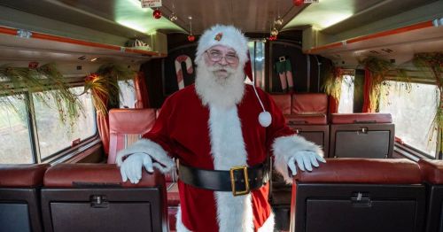 Santa Claus stands inside a decorated train car, wearing his traditional red suit and smiling warmly.