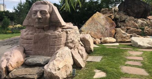 A stone sculpture of a sphinx surrounded by rocks and greenery in a landscaped area.