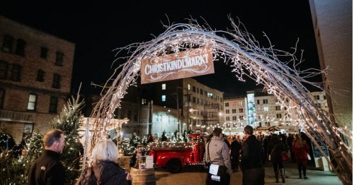 A festive market entrance adorned with lights, featuring a sign that reads "Christkindlmarkt" and a vintage red truck.