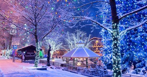 A snowy street scene adorned with colorful holiday lights and a gazebo surrounded by trees, creating a festive atmosphere.