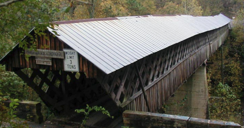 A wooden covered bridge with a sloped roof, surrounded by trees, featuring a weight limit sign.