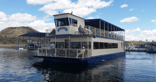 A large boat with a blue roof and upper deck, cruising on a lake with mountains in the background and people on board.