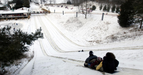 A snowy hill with people sledding down, surrounded by trees and a lodge in the background.