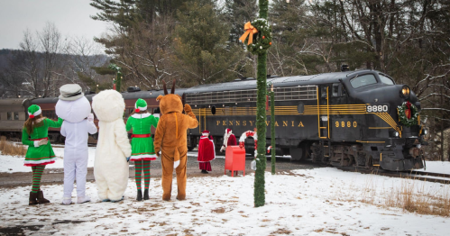 A festive scene with costumed characters watching a decorated train in a snowy landscape.