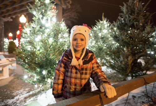 A smiling girl in a winter coat and Hello Kitty hat stands by decorated Christmas trees with lights.