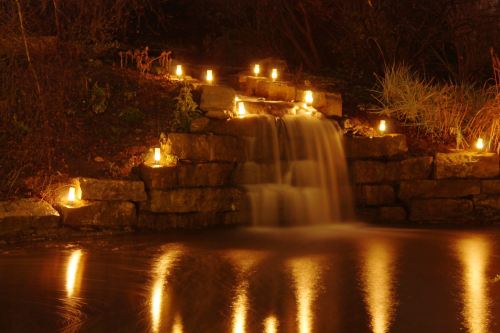 A serene waterfall at night, illuminated by glowing candles on the rocks, reflecting softly in the water below.