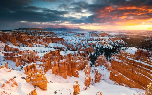 A stunning winter landscape of Bryce Canyon, featuring snow-covered orange rock formations under a dramatic sunset sky.
