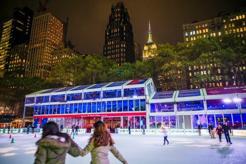 A nighttime ice skating rink in a city, with skyscrapers and the Empire State Building illuminated in the background.