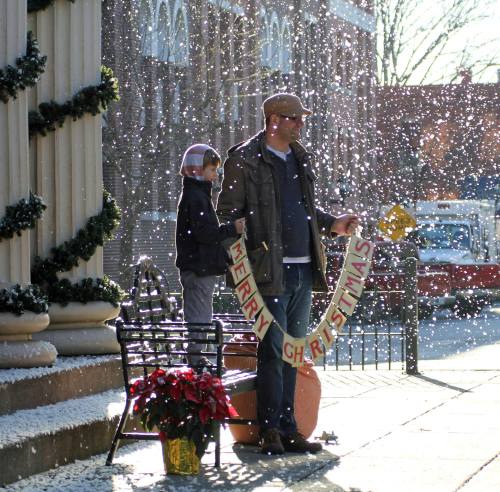 A man and a child stand outside, holding a "Merry Christmas" banner as snow falls around them.