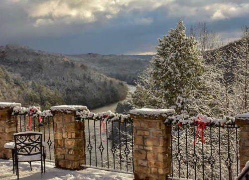 Snow-covered landscape with a river, trees, and a stone railing adorned with festive decorations.