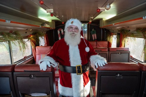Santa Claus in a red suit stands inside a decorated bus, smiling with festive ornaments and greenery around him.