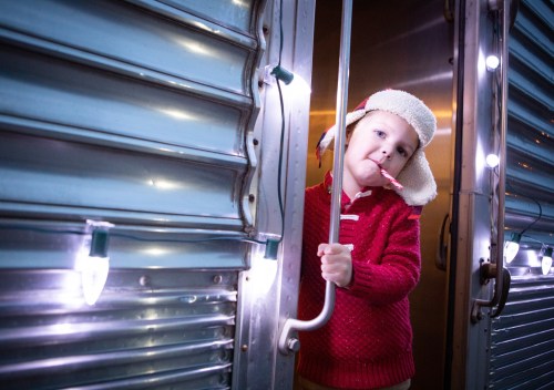 A child in a cozy red sweater and hat leans against a metal door, holding a candy cane, surrounded by soft lights.