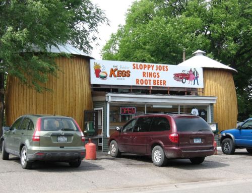 A rustic restaurant with a round wooden exterior, featuring a sign for Sloppy Joes, rings, and root beer. Cars parked outside.