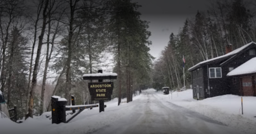 Snowy road leading to Aroostook State Park, flanked by trees and a rustic building on a winter day.