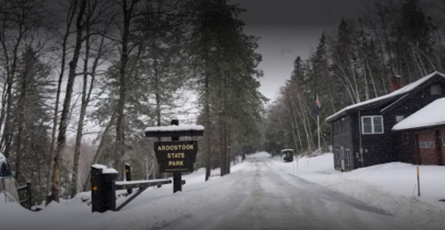 Snowy road leading to Aroostook State Park, with trees lining the path and a house on the right.