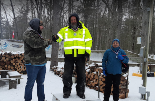 Three people stand on a podium in a snowy outdoor setting, with one wearing a bright yellow jacket and medals.