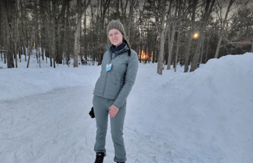 A person in winter attire stands on a snowy path surrounded by trees, smiling at the camera.