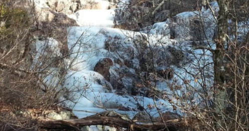 A frozen waterfall cascading over rocks, surrounded by bare trees and winter scenery.