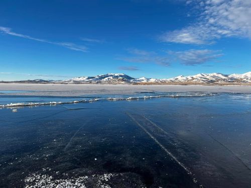 A clear, icy lake reflects the blue sky, with snow-capped mountains in the background under a few scattered clouds.