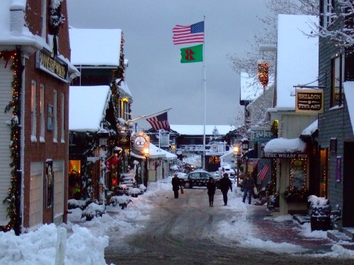 Snow-covered street lined with festive shops, American flag flying, and people walking in a winter scene.