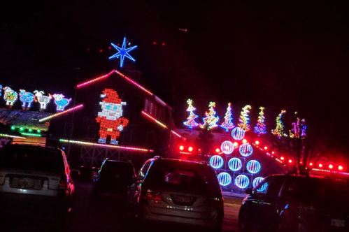 Colorful holiday lights decorate a house and trees, featuring a large Santa figure, with cars parked in the foreground.