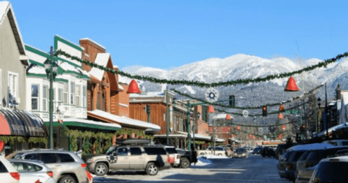 Snowy street scene decorated for the holidays, with mountains in the background and festive lights hanging above.