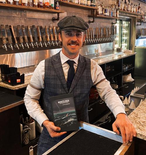 A man in a vest and cap stands at a bar, holding a book titled "Money's Back." Behind him are beer taps and bottles.