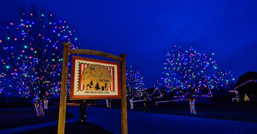 A festive scene with colorful Christmas lights on trees and a sign for "The Christmas Factory" against a twilight sky.