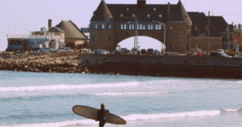 A surfer walks towards the ocean with a surfboard, with a historic building in the background.