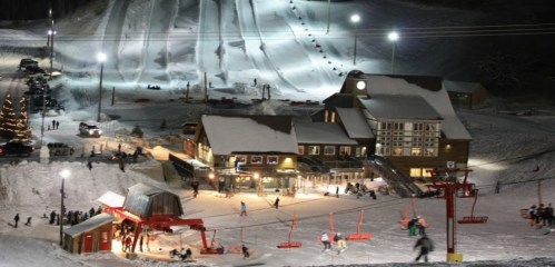 A snowy ski resort at night, featuring a lodge, ski lifts, and people skiing and snowboarding on illuminated slopes.