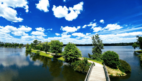 A serene lake scene with lush greenery, a wooden walkway, and a bright blue sky filled with fluffy clouds.
