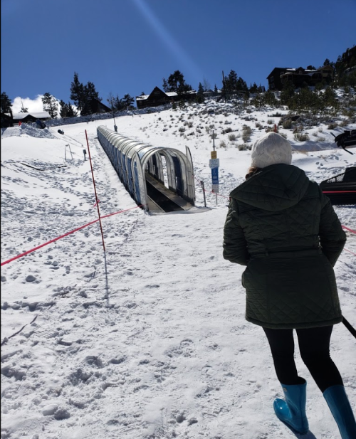 A person in winter attire walks towards a snow-covered slope with a ski lift in the background under a clear blue sky.