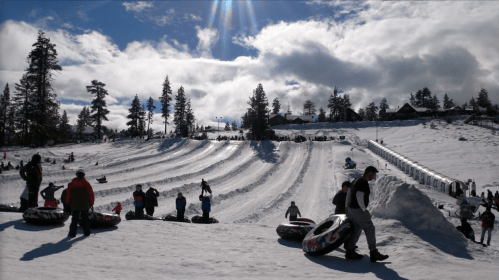 A snowy landscape with people tubing down slopes, surrounded by trees and a bright blue sky with clouds.