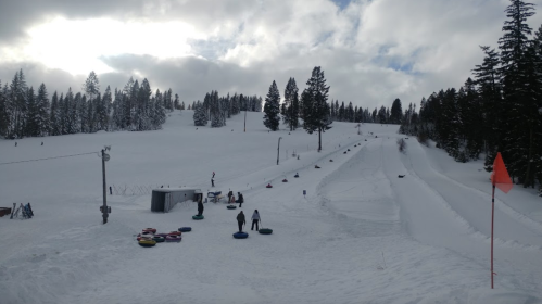 A snowy landscape with people tubing down a hill, surrounded by trees and a cloudy sky.